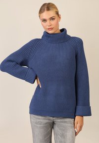 Blauer Strickpullover mit hohem, geripptem Kragen und breiten Bündchen. Die Struktur ist dick und hat ein vertikales Muster. Getragen mit grauen Jeans.