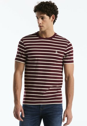 Junger Mann trägt ein kurzärmeliges T-Shirt mit bordeaux-weißen Streifen und blaue Jeans, schaut nach links.