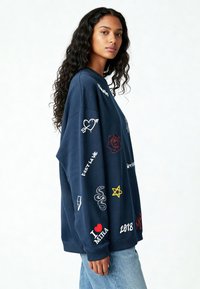 Marineblauwe sweatshirt met een oversized pasvorm, voorzien van kleurrijke geborduurde patches, waaronder harten, een roos, slangen, bliksem en tekstaccenten.