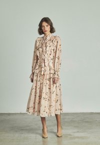 Vestido largo y fluido hecho de tela de color rosa pálido con un estampado floral. Presenta volantes, un cuello con lazo y mangas largas. Zapatos de tacón beige acompañan el conjunto.