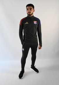 Chándal atlético negro con cuello con cremallera, rayas rojas y azules en las mangas, detalles dorados, logo en el pecho y pantalones negros ajustados. Zapatillas negras.