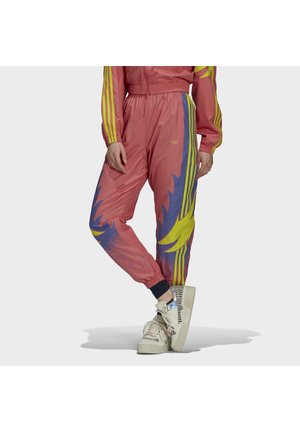 Personne portant un pantalon de sport rose avec des motifs de flammes jaunes et violettes et des manches rayées jaunes, debout jambes croisées et baskets blanches.