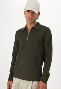 Olive green, long-sleeve pullover with a zippered collar, made from a soft material, worn with light-colored trousers and a beige cap.