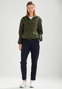 Olivenfarget, lett jakke med glidelås og hette, frontlomme, kombinert med marineblå stripete bukser og hvite joggesko.