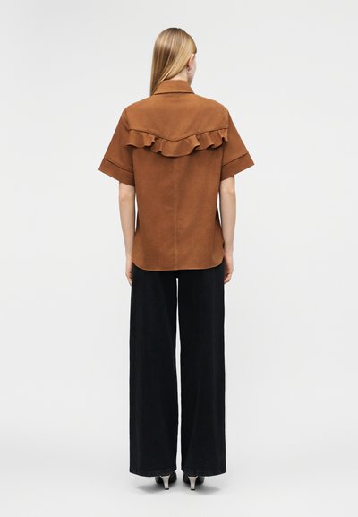 Mujer de pie, de espaldas, vistiendo una camisa marrón de manga corta con detalles de volantes y pantalones anchos negros, sobre un fondo gris.