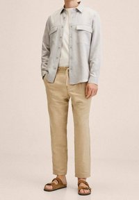 Camisa gris claro abotonada con dos bolsillos en el pecho, camiseta crema, pantalones beige y sandalias marrones. Tejido suave con ajuste relajado.