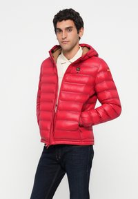Man wearing red puffer jacket with hood over beige polo shirt and dark jeans, standing with hands in jacket pockets against white background.