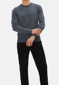 Pull gris tricoté avec un col rond, des manches longues et un ourlet côtelé, associé à un pantalon noir. Texture subtile sur tout le tissu.