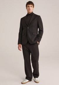 Schwarzer Anzug mit einem einzigen Knopf und zwei Fronttaschen, kombiniert mit einem passenden Rollkragenpullover und lässigen Sneakern in Beige und Weiß.