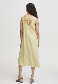 Rochie midi galbenă din in, cu decolteu rotund și spate deschis, având un sistem de închidere cu fundă. Purtați cu mules cu toc transparent.