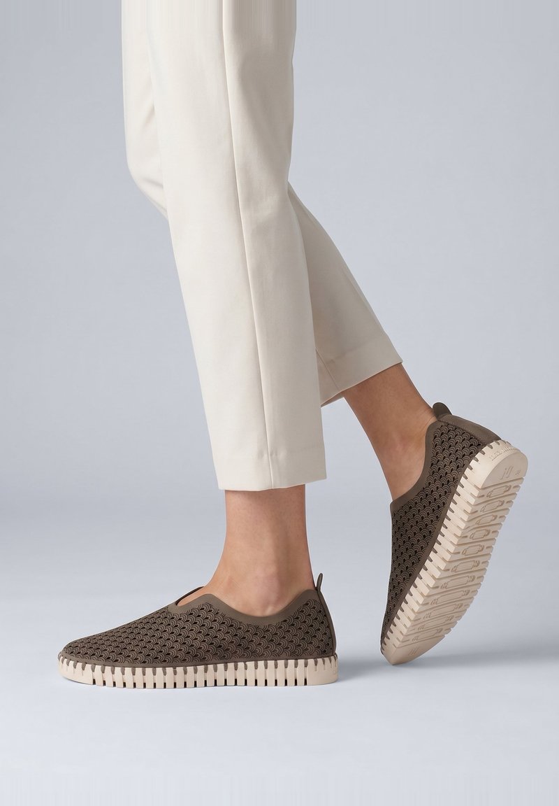 Person iført beige bukser og mørkebrune slip-on sko med tekstureret sål mod en ensartet lys grå baggrund.