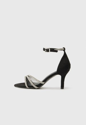 Black heeled sandal with a suede texture, featuring crisscross mesh straps adorned with silver accents and an ankle buckle.