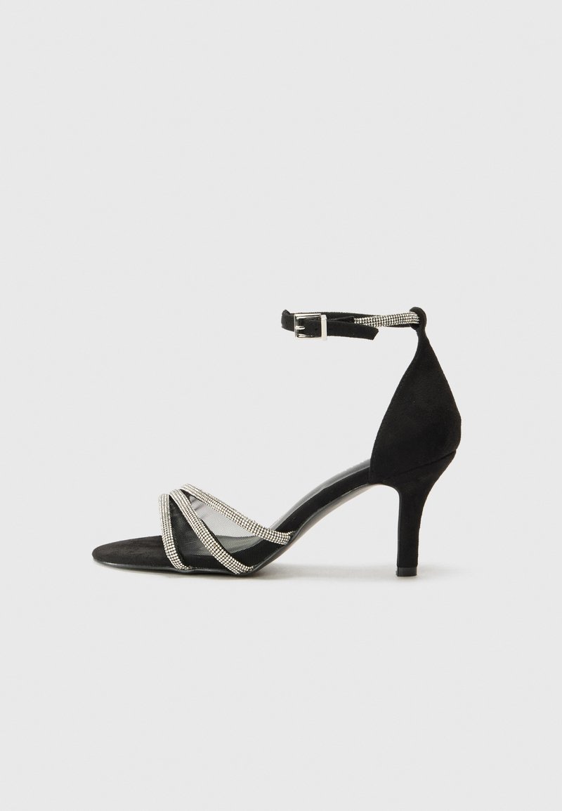 Schwarze High Heel Sandale mit Wildlederoptik, ausgestattet mit überkreuzten Netzzurriemen, die mit silbernen Akzenten verziert sind, sowie einer Schnalle am Knöchel.