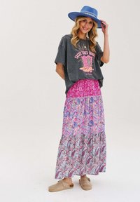 Sivý grafický tričko s růžovým potiskem, přehozené přes růžovou maxisukni s paisley vzorem. Modrý široký klobouk a béžové nazouvací boty.
