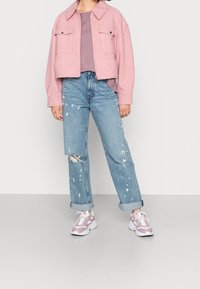 Rosa kurze Jacke mit zwei Vorderentaschen, kombiniert mit blauen, ausgewaschenen Jeans, die Farbspritzer aufweisen, und hellrosa Sportschuhen.