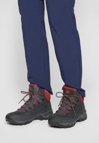 Scarpe da trekking nere con accenti rossi, caratterizzate da una tomaia testurizzata, colletto imbottito e suola resistente. I lacci di un colore contrastante migliorano il design.
