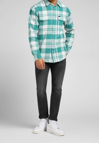 Chemise en flanelle à carreaux verts et blancs avec manches longues, boutonnière avant, deux poches poitrine, portée avec un jean noir et des baskets blanches.