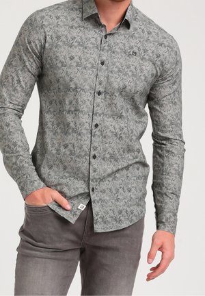 Mann trägt ein graues, gemustertes, langärmliges Hemd mit dunklen Knöpfen und dunkelgraue Hose, Hand in der Hosentasche, vor einfachem Hintergrund.