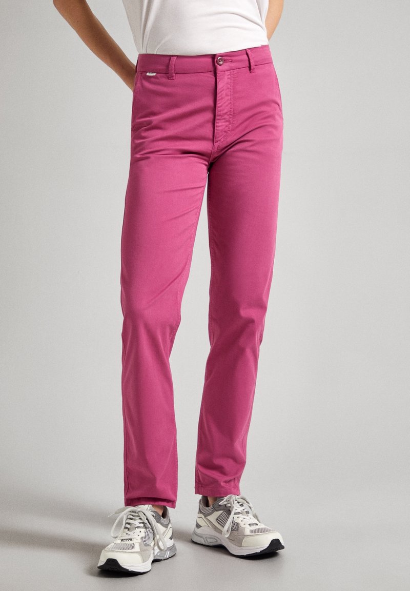 Chinos en coton rose avec une coupe slim, dotés d'une fermeture à bouton et de poches, associés à des baskets grises et blanches. Texture lisse.
