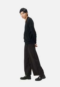 Svart stickad kofta med v-ringning och fickor, kombinerad med vida svarta byxor och svarta ankelboots, poserad mot en neutral bakgrund.