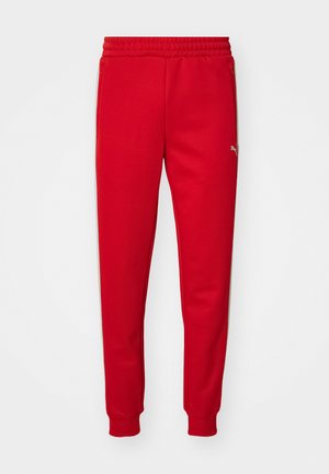 Red sweatpants made of soft fabric with an elastic waistband, tapered legs, and gold logo detail on the left side. Smooth texture.