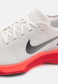 Fehér sportcipő textúrázott felsőrésszel, fekete Nike logóval és élénkpiros talppal. Laposszemély zapfapántokkal és párnázott bokarésszel rendelkezik.