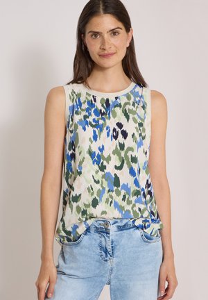 Femme aux longs cheveux foncés portant un haut sans manches à motifs floraux et un jean en denim bleu clair, debout devant un fond uni.