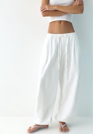 Person iført løse hvide bukser, hvid cropped tanktop og lysegrå klip-klapper, stående med armene krydset mod en hvid baggrund.