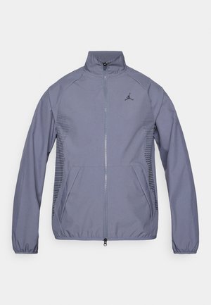 Ljusgrå zip-up jacka med hög krage, sidofickor, mesh-detaljer och en svart logotyp på bröstet. Slät, atletisk tyg.