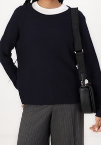 Person, der einen marineblauen Pullover über einem weißen Hemd, graue Nadelstreifenhose trägt und eine schwarze Crossbody-Tasche mit breitem Tragegurt über der Schulter trägt.