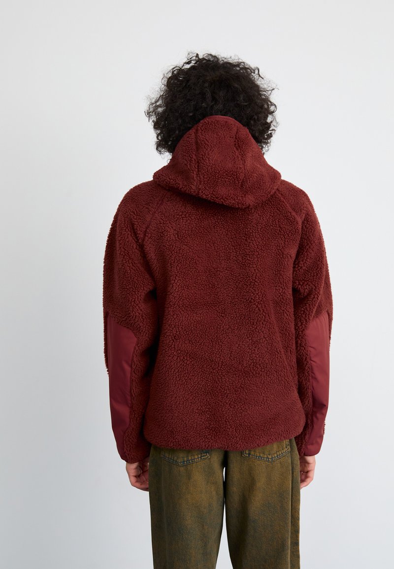 Veste en polaire bordeaux avec un matériau texturé et duveteux, une capuche et des panneaux en tissu bordeaux contrastants sur les manches.