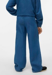 Modri jeans širokih hlač z elastiko v pasu, ki imajo gladko teksturo in subtilne detajle šivanja, v kombinaciji z črnimi čevlji.