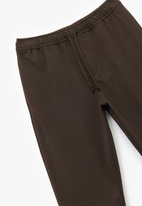 Jogging en brun, fabriqué en tissu texturé, avec une taille élastique et un cordon de serrage, et une coupe fuselée. Pas de poches visibles.