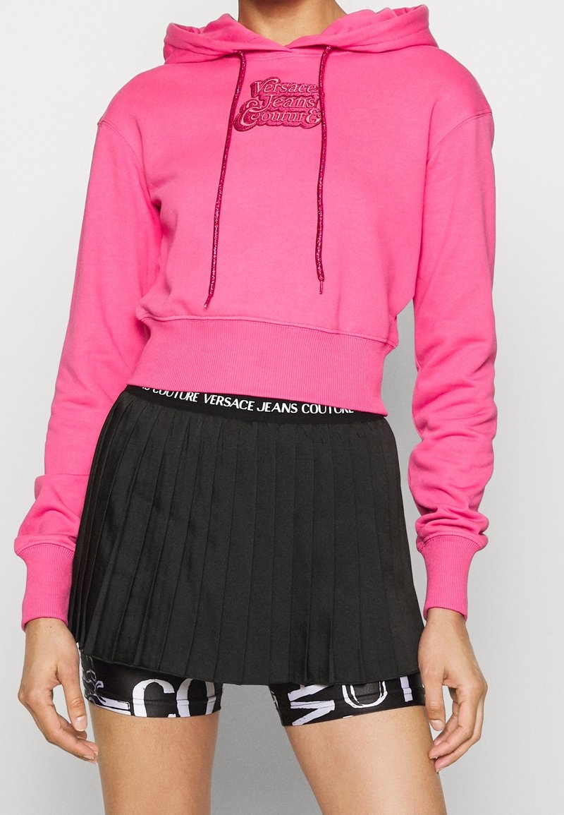 Femme portant un sweat à capuche court rose vif de Versace Jeans Couture, une jupe plissée noire et un short noir à motifs en dessous.
