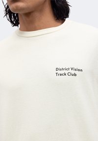 Κοντινό πλάνο ενός ατόμου που φοράει λευκό T-shirt με το κείμενο "District Vision Track Club" τυπωμένο σε μαύρο στο αριστερό στήθος.