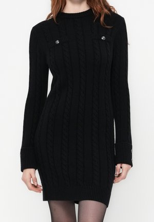 Femme portant une robe-pull en maille torsadée noire avec des poignets boutonnés et des détails décoratifs de boutons près de la poitrine.