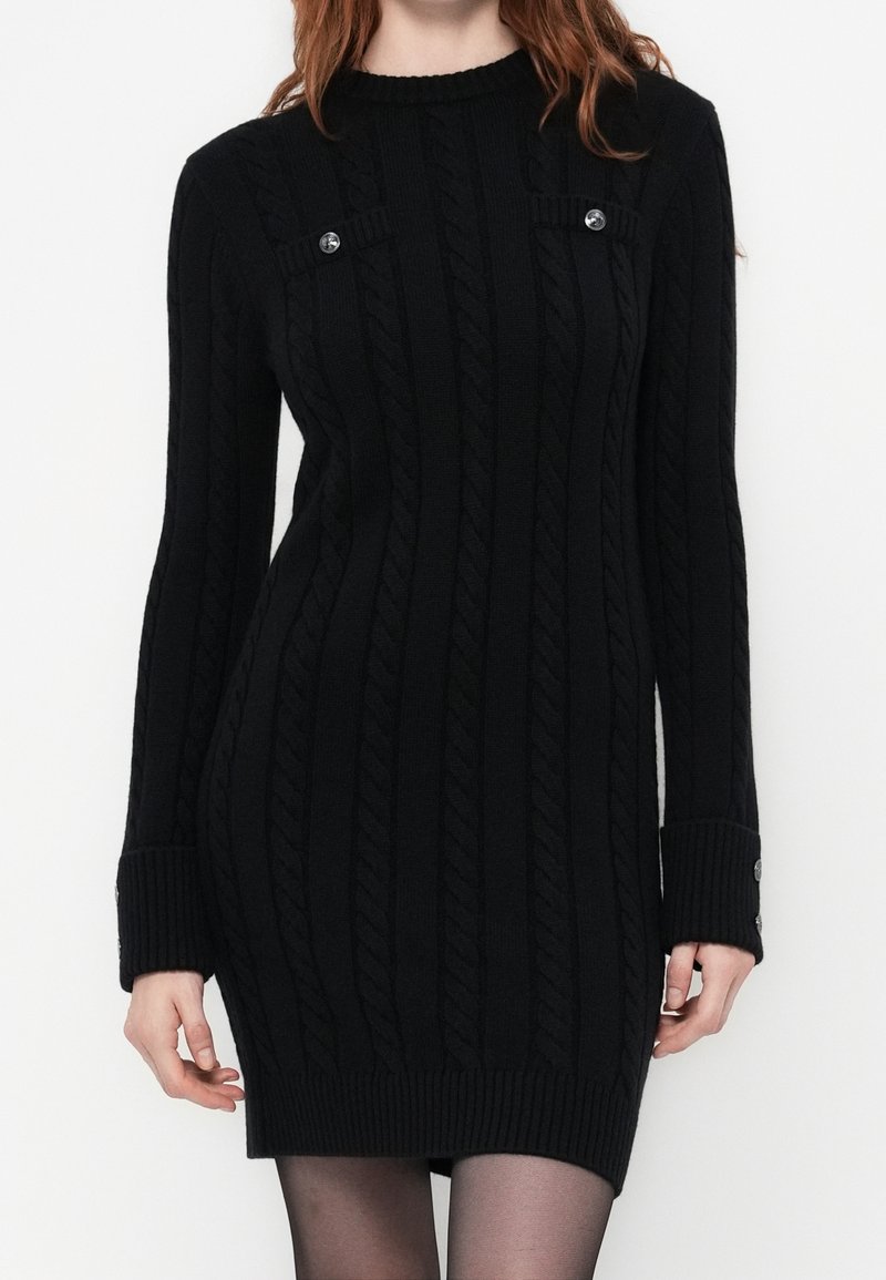 Femme portant une robe-pull en maille torsadée noire avec des poignets boutonnés et des détails décoratifs de boutons près de la poitrine.