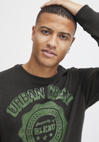 Sudadera negra con un diseño gráfico verde que incluye el texto "URBAN CREW" y un logo circular. Tejido suave y casual; con cuello redondo.
