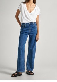 Žena v bílém tričku s výstřihem do V, v modrých rovně střižených džínách s vysokým pasem a v černých sandálech na otevřené špičce stojí s jednou rukou za zády.