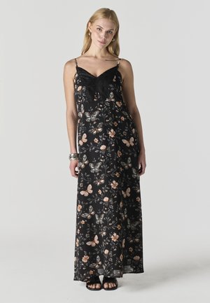 Mujer vistiendo un vestido largo negro con estampado floral, detalle de encaje y tirantes finos, de pie frente a un fondo liso gris claro.