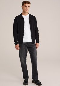 Zwarte geribbelde cardigan met ritsaanluiting, gedragen over een wit t-shirt; gecombineerd met donkere jeans en zwarte schoenen. Effen achtergrond.