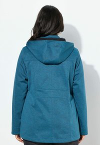 Nainen, jolla on pitkät tummat hiukset, pukeutuneena siniseen huppariin, kääntynyt pois yksinkertaista valkoista taustaa vasten.