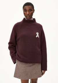 Bordeaux trui met hoge kraag, zachte textuur en wit embleem op de borst. Gepaard met een bruine rok met een subtiel patroon.