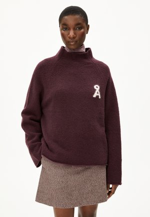 Pull bordeaux à col montant, en tissu doux et texturé, avec un emblème blanc sur la poitrine. Associé à une jupe marron avec un motif subtil.