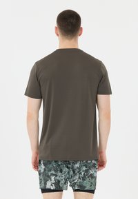 Mies, joka katsoo poispäin, päällään yksinkertainen tummanruskea t-paita ja musta-vihreä kuvioitu shortsit valkoista taustaa vasten.