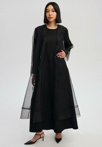 Must kleit läbipaistvate pikkade varrukatega ja põrandapikkusega disainiga. Omab istuvat ülakeha ja voolavat kihilist siluetti. Õhukesed kontsaga kingad.