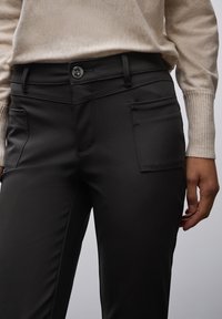 Osoba nosząca czarne dopasowane spodnie z przednimi kieszeniami naszytymi oraz beżowy sweter z długim rękawem w prążek, ręka spoczywająca na boku.