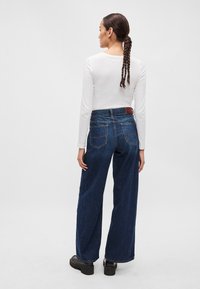 Jeans de denim de perna larga em azul escuro com uma cintura alta, apresentando bolsos traseiros e um patch de pele na parte de trás. Combinado com uma blusa de manga longa branca.