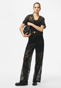 Črn čipkast top in hlače z rožnatimi vzorci in sequini. Model drži majhno črno torbico, nosi ravne čevlje in minimalističen dizajn.