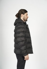 Svart puffjacka med huva, med en quiltad design, glänsande yta, framknappar och elastiska ärmslut. Bärs över svarta byxor.